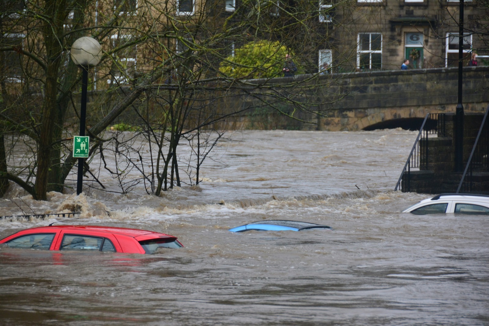 Three cars partially submerged in a flooded river near a stone bridge