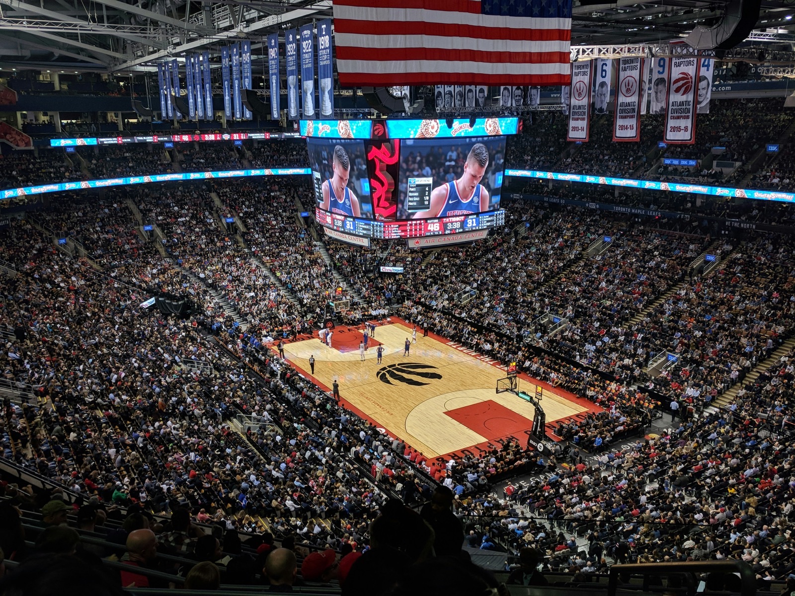 A basketball arena exterior at dusk with illuminated signage and a small crowd of fans walking toward the entrance.